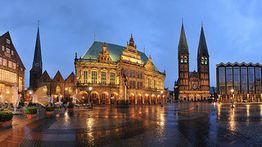 Bremer Rathaus und Dom bei Abenddämmerung, beleuchtet, mit nassem Kopfsteinpflaster im Vordergrund.