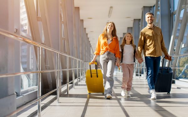Vater, Mutter und Kind laufen mit Koffern in einem Flughafen entlang. 
