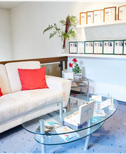 Heller Warteraum mit weißem Sofa, roten Kissen, Glastisch, roten Schränken und Pflanzen.