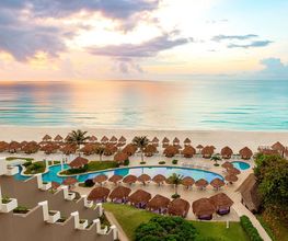 Playa al amanecer con palapas, piscina y vista al mar desde un hotel.