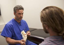 Doctor in blue scrubs explaining spine model to a seated patient.