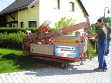 Mann bedient Bohrmaschine im Garten vor gelbem Haus. Schild: "Brunnenbau & Bohrservice".