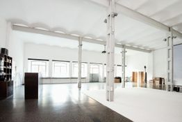 Helles, industrielles Loft mit großen Fenstern, Bücherregal und weißem Bodenbereich.