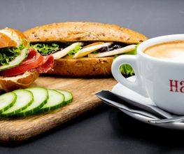 Brötchen mit Wurst und Käse, daneben eine Cappuccino-Tasse auf einem Tablett, Gurkenscheiben vorne.