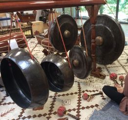 Mehrere große Gongs hängen an einem Holzgestell, umgeben von Teppichen und Musikinstrumenten.