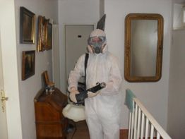 Person in Schutzanzug und Maske mit Gerät in einem Flur stehend.