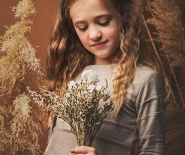 Niña con lazo en el cabello sostiene flores y mira hacia abajo, rodeada de plantas secas.