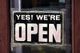 Schild an Tür mit der Aufschrift "YES! WE'RE OPEN" in schwarz-weißem Design.
