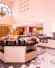 Restaurante amplio con bufé variado y mesas preparadas bajo techos altos y ventanas grandes.