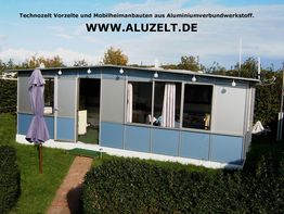 Ein Mobilheim mit Aluminiumverkleidung, daneben ein Sonnenschirm; umgeben von Hecken.