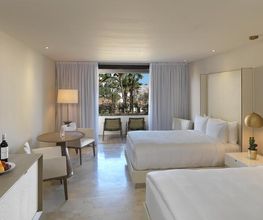 Habitación de hotel moderna con dos camas, balcón, lámpara y vista a palmeras afuera.