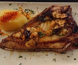 Pescado asado con ajo en rodajas y papa hervida espolvoreada con pimentón, servido en un plato blanco.