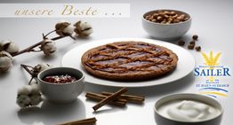 Ein runder Kuchen mit Marmeladenfüllung, Zimtstangen und Schalen mit Sahne und Nüssen auf dem Tisch.