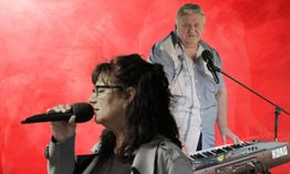 Zwei Personen mit Mikrofon und Keyboard auf rotem Hintergrund. Eine singt, der andere spielt Keyboard.