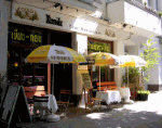 Straßencafé mit gelben Sonnenschirmen und Holztischen auf Gehweg unter Sonnenlicht.