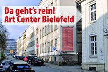 Straße mit Gebäuden, Schild: "Da geht's rein! Art Center Bielefeld", Autos am Straßenrand.