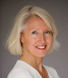 Eine lächelnde Frau mit blondem, schulterlangem Haar vor grauem Hintergrund.