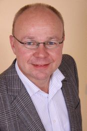 Mann mit Brille, lächelnd, gestreiftes Hemd und kariertes Sakko, beige Hintergrund.