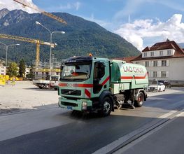 Straßenkehrmaschine fährt auf Straße vor Baugelände und Bergen im Hintergrund.