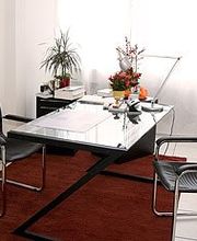 Minimalistisches Büro mit Glas-Schreibtisch, zwei schwarzen Stühlen und Pflanzen auf rotem Teppich.