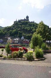 Rote Autos vor einer Burg auf einem grünen Hügel, mit bunten Häusern im Vordergrund.