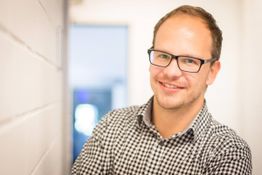 Mann mit Brille und kariertem Hemd lächelt in die Kamera vor unscharfem Hintergrund.