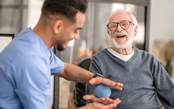 Pfleger kümmert sich mithilfe eines hellblauen Balles mit Noppen um die Motorik seines Patienten.