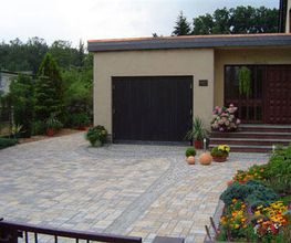 Einfahrt mit gepflastertem Boden, Garage, Topfpflanzen und Blumenbeeten vor einem Haus.