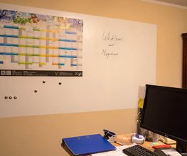 Whiteboard mit Magneten, links Wandkalender. Vorne Schreibtisch mit Monitor und Ordner.