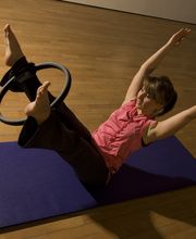 Person macht Pilates auf Matte, nutzt Fitnessring mit Füßen, hebt Arme in hellem Raum.