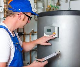 Techniker in blauer Latzhose prüft eine Heizungsanlage und notiert Messwerte auf einem Klemmbrett.