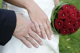 Hände eines frisch verheirateten Paars mit Eheringen und Brautstrauß aus roten Rosen.