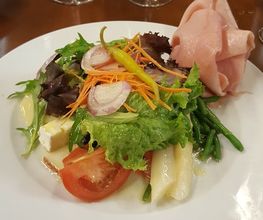 Plato de ensalada variada con jamón y vegetales, al fondo copas de vino tinto.