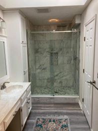 White bathroom with a glass shower, marble tiles, wooden floor, and a colorful rug.