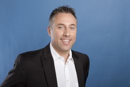 Ein lächelnder Mann im Anzug vor einem blauen Hintergrund.
