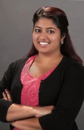 Smiling person with long hair, wearing a pink top and black sweater, arms crossed in front.