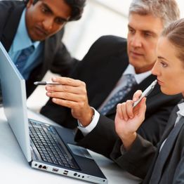 Three professionals in suits discuss at a laptop, pointing and making notes.