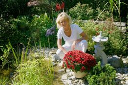 Person in weißer Kleidung kniet im Garten neben roten Blumen und einem kleinen Steinturm.