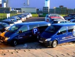 Zwei blaue Firmenvans geparkt neben vielen Autos auf einem Parkplatz vor einem Industriegebäude.