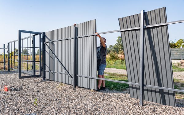 Instalación de valla metálica en terreno con ligera pendiente, con un operario ajustando paneles de chapa sobre estructura de acero en exterior.