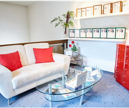Heller Warteraum mit weißem Sofa, roten Kissen, Glastisch, roten Schränken und Pflanzen.