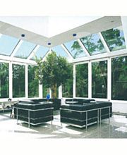 Moderner Wintergarten mit Glasdach, schwarzen Sofas und großem Baum, viel Tageslicht.