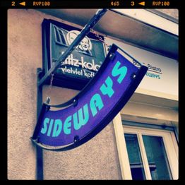 Blaues Schild mit der Aufschrift "SIDEWAYS" hängt schräg an einer Wand über einem Fenster.