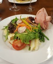 Plato de ensalada variada con jamón y vegetales, al fondo copas de vino tinto.