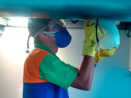 Técnico de máscara e luvas realiza manutenção em equipamento sob mesa iluminada.