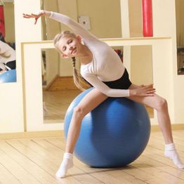 Girl balancing on a blue exercise ball, wearing a white top and black shorts in a dance studio.