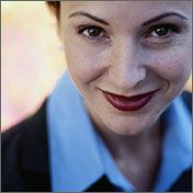 Frau lächelt direkt in die Kamera, trägt Blazer und Hemd. Hintergrund unscharf.