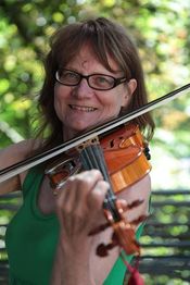 Frau spielt Geige im Freien, trägt Brille und grünes Oberteil, lächelnd mit unscharfem Hintergrund.