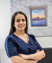 Mujer con uniforme azul, brazos cruzados, frente a cuadro de paisaje de mar y cielo.
