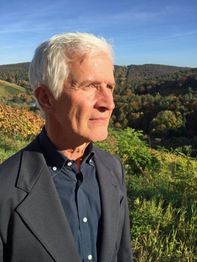 Ein älterer Mann steht im Freien mit hügeliger Landschaft im Hintergrund, der Blick in die Ferne.